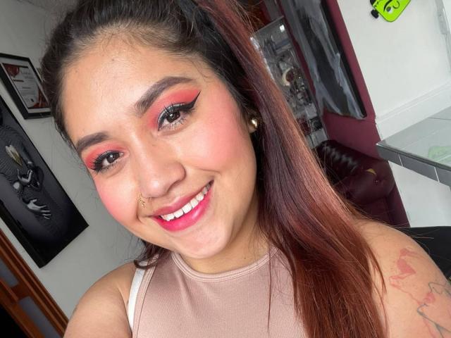 ValmontAlessia with red and pink eye makeup, smiling, brown hair, nose ring, pink lipstick, tattoo on right arm, wearing beige top, in selfie, showing white teeth.