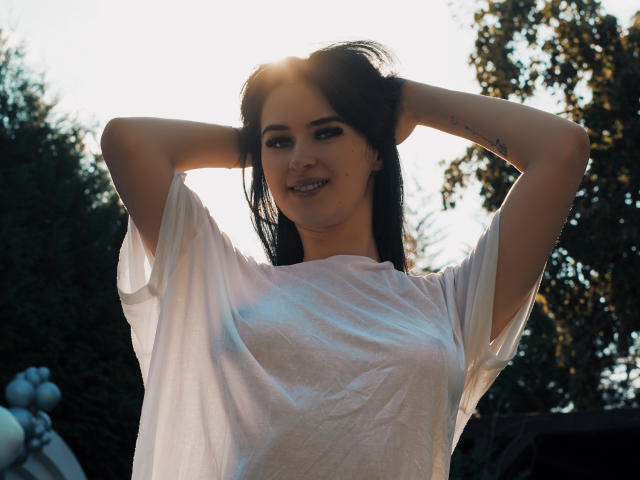 EvelinSkye with hands in dark hair, wearing a white, slightly transparent shirt, sun in background, smiling, small tattoo on right forearm, visible nipples through shirt.