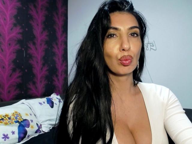 BrunetteBabe69 with long black hair, pouty red lips, and large tits in a low-cut white top, making a kissing face in a selfie.