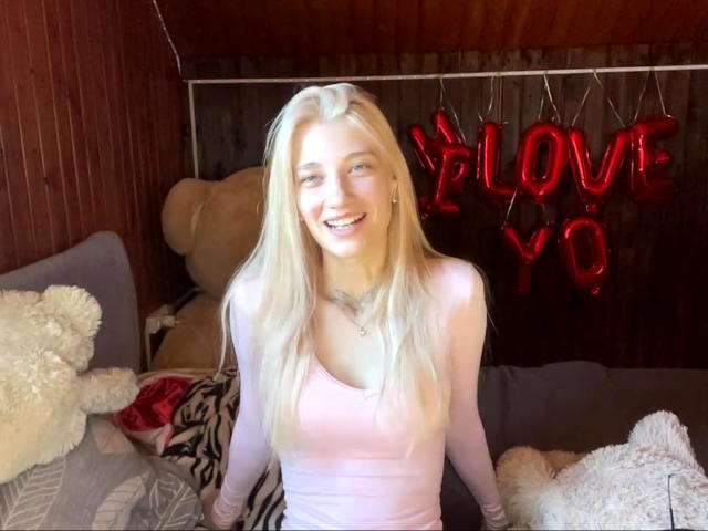 Georgann Whatcott, con i capelli biondi, indossa una maglietta rosa a maniche lunghe, sorride e siede sul letto con due orsacchiotti di peluche bianchi e grigi, un cartello luminoso rosso con la scritta "LOVE", un soffitto in legno e una coperta a motivi zebrati..