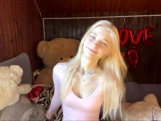 GeorgannWhatcott with long blonde hair, pink tank top, and tattoo on chest, smiling, sitting in bed with large teddy bear and red heart-shaped balloon.