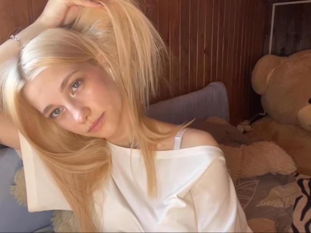 Georgann Whatcott, con i capelli biondi raccolti in due codini, indossa una camicia bianca con una spalla scoperta, guarda la fotocamera con gli occhi verdi e siede accanto a un grande orsacchiotto di peluche..