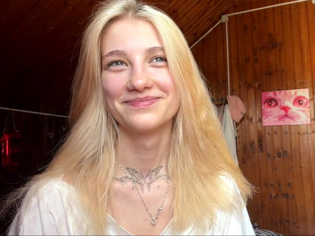 GeorgannWhatcott with blonde hair, blue eyes, and a tattoo under her neck, wearing a white top, smiling slightly. Wooden wall and pink cat painting in background.
