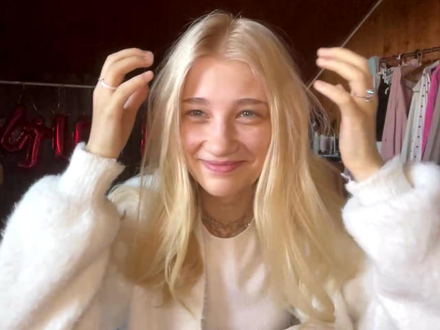 Georgann Whatcott, con i capelli biondi, sorridente, indossa un maglione bianco e morbido, tiene i capelli con entrambe le mani, porta una collana e degli anelli, in un ambiente interno accogliente e ben illuminato..