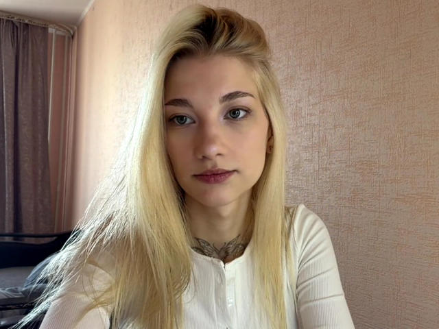Georgann Whatcott ha lunghi capelli biondi, occhi verdi e un piccolo tatuaggio sul collo, indossa una camicia bianca e ha un'espressione neutra in una stanza molto luminosa..