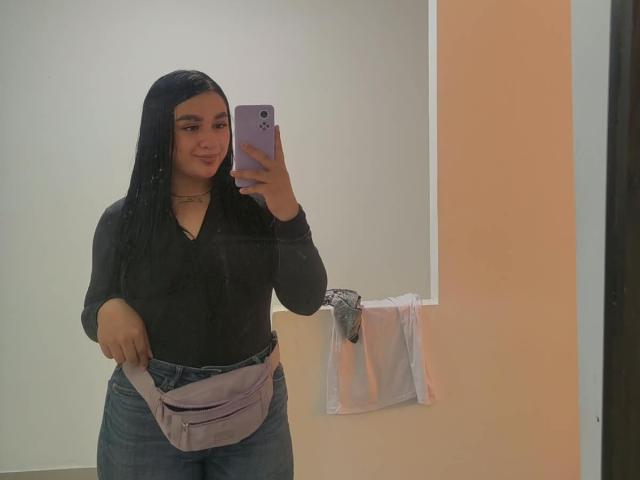 SamyBennet, avec de longs cheveux noirs, portant une chemise noire, un jean bleu et une ceinture violette, tenant un téléphone violet, souriant dans un selfie pris dans un miroir..