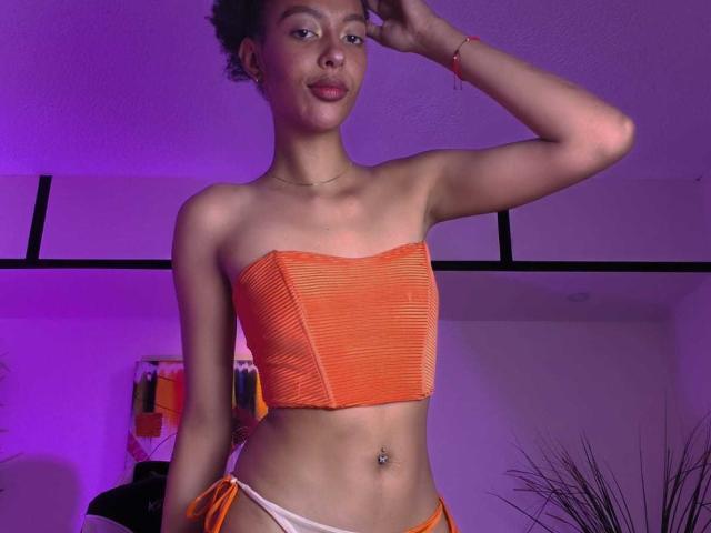 KiaraKamau, mit dunkler Haut und kurzen, lockigen Haaren, trägt ein orangefarbenes, geripptes Top und einen weißen Bikini-Unterteil mit seitlichen Bändern. Sie posiert mit einer Hand auf dem Kopf, das Licht ist violett..
