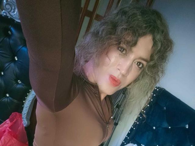 RossSofiia, met krullend grijs haar, een bruine top en rode lippenstift, maakt een selfie en laat haar borsten zien, terwijl ze staat voor een zwart hoofdeinde met een pluizige, gestikte bekleding..