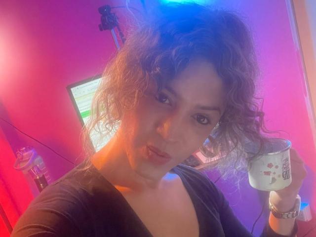 RossSofiia, com cabelos castanhos cacheados, segurando uma caneca branca com desenhos vermelhos, vestindo uma blusa preta, em um quarto iluminado em tons de rosa e azul, olhando para a câmera..