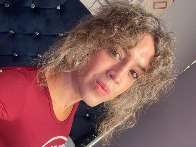 RossSofiia mit lockigem, graubraunem Haar, rotem Hemd, einem gepolsterten, dunkelblauen Sessel, leicht gerümpften Lippen, die zur Seite schaut, in einem Selfie..