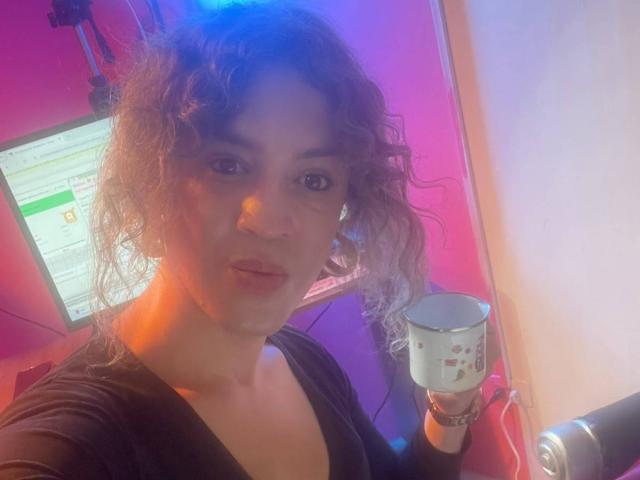 RossSofiia, con cabello rizado, sosteniendo una taza, vistiendo una blusa negra, mirando a la cámara, luces rosas y azules, pared blanca en el fondo, maquillaje sutil, labios ligeramente fruncidos..