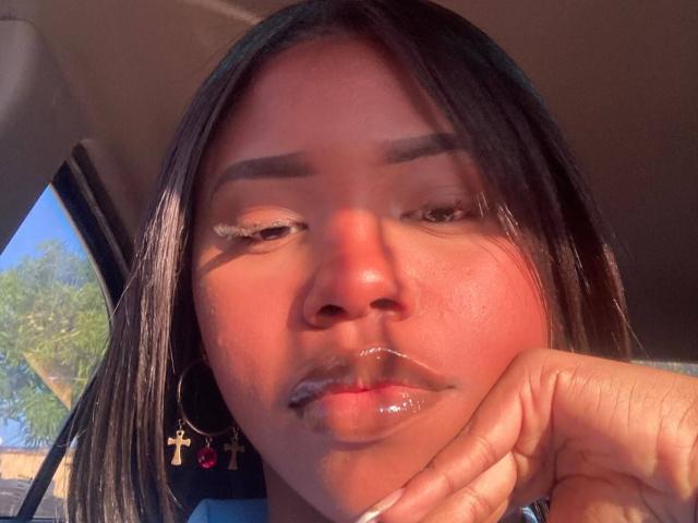 CatalinaSanA with shiny lips, dark hair, and gold cross earrings, resting her hand on her cheek, looking down with a slight pout, sunlight highlighting her face.