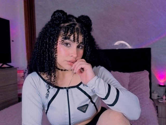 VanessaBlake with curly black hair in double buns, wearing a gray long-sleeve top, black choker, and black shorts, sitting on a bed with pink pillows, hand on chin.