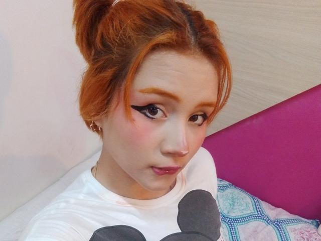 ChloeeWilliams with orange hair, black winged eyeliner, pink blush, white shirt with gray polka dots, looking at camera, pink headrest, and blue patterned pillow in background.