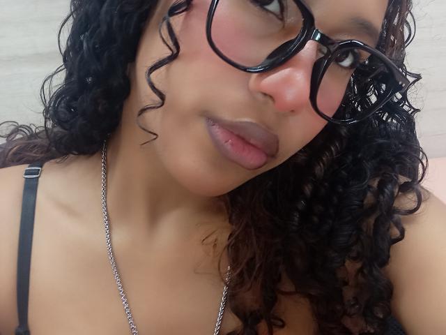 LovelyConnie with curly black hair, wearing black glasses and a black strap top, has a cute face, pinkish cheeks, and slightly parted pinkish lips. Necklace visible.