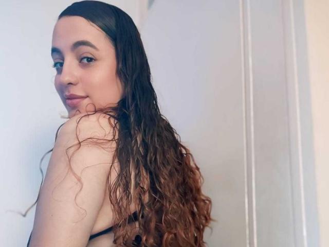 StacyMillerr with long, wet brown curly hair, looking over shoulder, showing bare back and small tits in black bra, light skin, subtle smile, soft makeup, indoors.