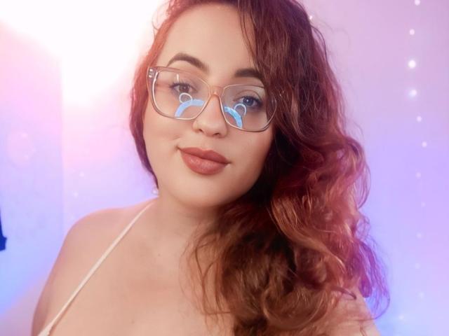 StacyMillerr with wavy red hair, wearing clear glasses and a white halter top, showing her cute face and pouty lips. Her tits are small and she's smiling.