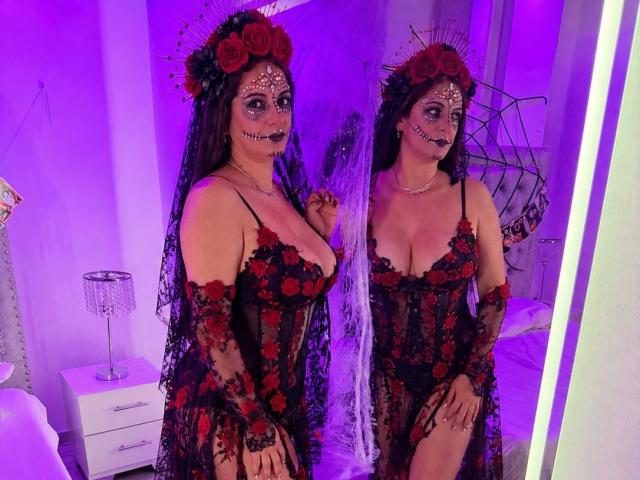 MadameScarlett in a black lace dress with red roses, flower headpiece, and skull makeup, showing off her big tits and reflecting in a mirror with purple lighting.