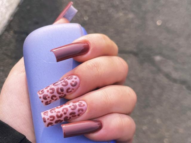 La mano de Tinelia sostiene una botella azul, mostrando uñas largas, brillantes y de color rosa dorado, con puntas estampadas con un diseño de leopardo en rosa. Su manicura, impresionante, es perfecta, amigo..