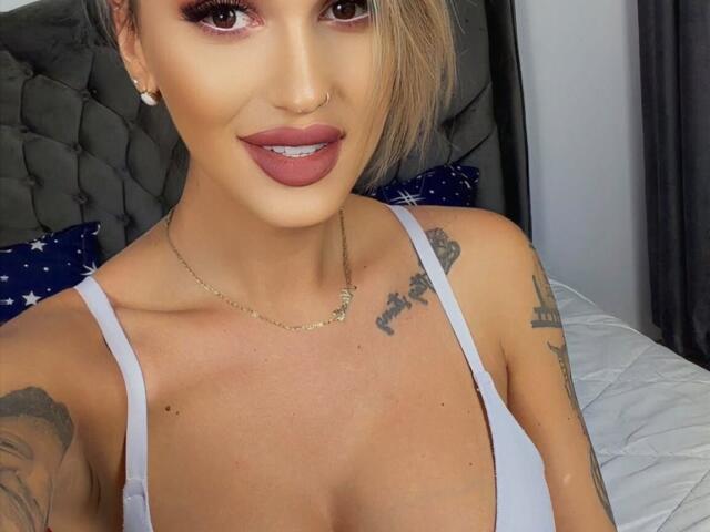 NicolSNaya with tan skin, blonde hair, and pink lipstick, wearing a white tank top, showing tattoos on her chest and arms, and a nose ring. Boobs visible, smiling.
