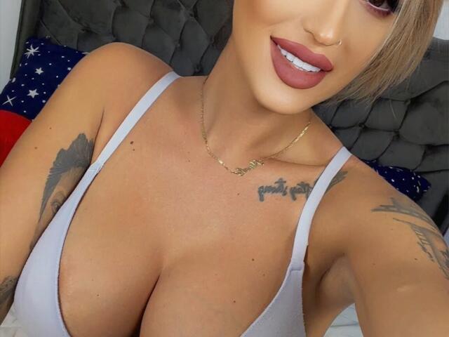 NicolSNaya with big tits, wearing a white bra, has tattoos on her arms and chest, a nose ring, and a gold necklace, smiling with red lipstick.