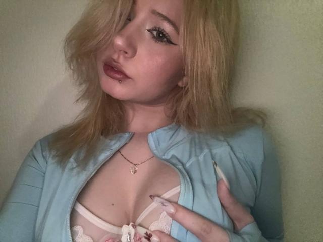 SerafinaDinwiddie with blonde hair, pouty lips, and a lip piercing, wearing a blue cardigan open to reveal white lace-up bra, touching her tits with a hand.