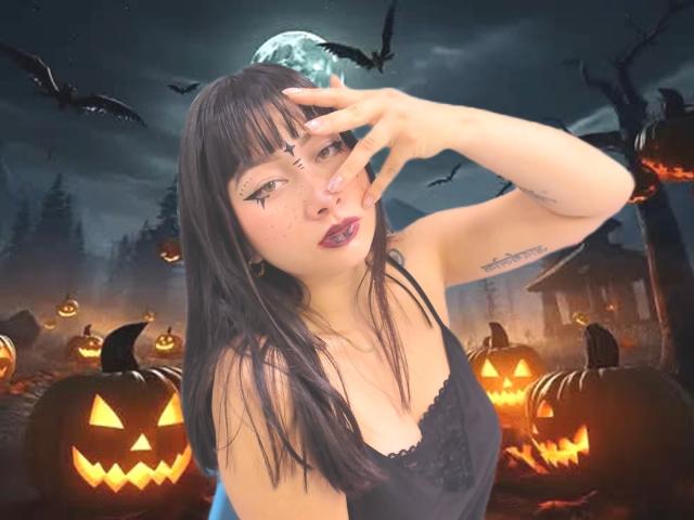 Genesiss with black hair, eyeliner, and red lipstick, wearing a black lace top, making a peace sign, surrounded by glowing jack-o'-lanterns, bats, and a full moon.