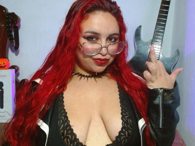 Estefy69 with red hair, glasses, black lace top, showing big boobs, spiked choker, and holding a guitar, smiling, middle finger up.
