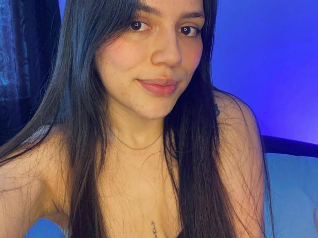 SophiaMarx with long black hair, light skin, small nose, and a slight smile, showing bare shoulders and a navel piercing, against a blue-lit background.