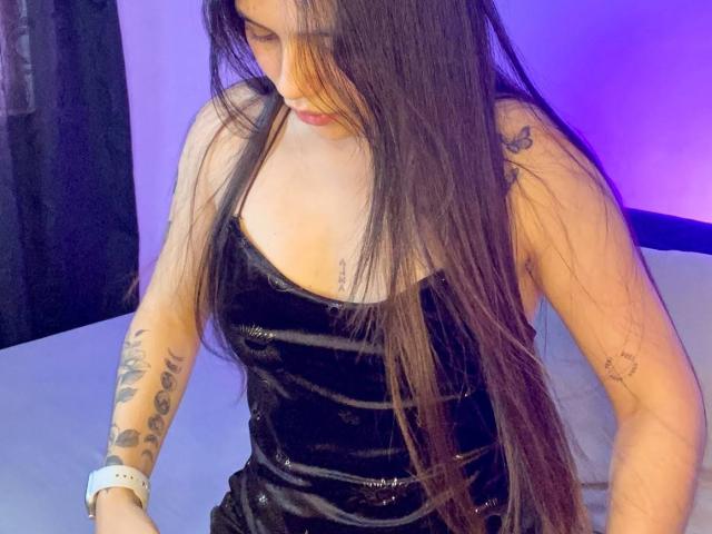 SophiaMarx with long brown hair, black velvet dress, tattoos on arms, white wristband, looking down, purple-blue light in background. Boobs visible under dress.