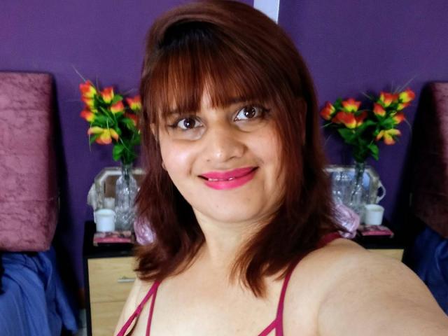VivianFranco with brown hair, bangs, pink lipstick, smiling, wearing a pink bra, holding a selfie, pink flowers in background, purple wall, blue bed, pink pillow.