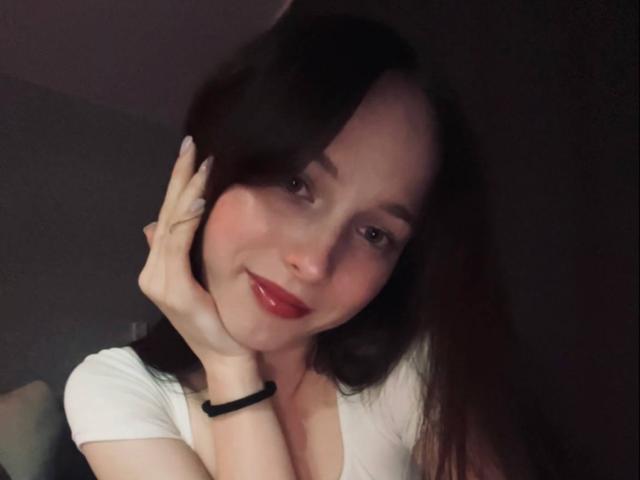 MercedezBanks, con capelli castano scuro, rossetto rosso e top bianco, sorridente, con una mano sul viso, che mostra piccoli seni, un braccialetto nero, illuminazione soffusa, un leggero rossore e un'espressione rilassata..
