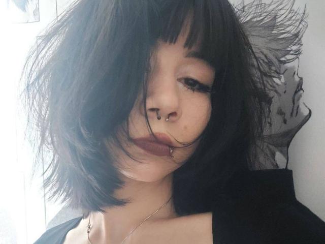 TempleWydner with messy black hair, nose piercings, lip ring, dark lipstick, and a black top, looking slightly to the side with a half-smile.