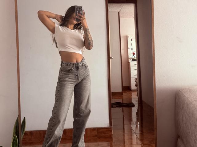 ThaliaAres with arms up, white crop top, gray high-waisted jeans, tattooed arm, holding phone, standing in room, reflecting in mirror, showing her ass and small tits.