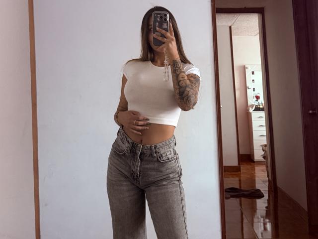 Thalia Ares, con cabello castaño largo, top blanco corto que deja ver pequeños senos, brazo tatuado, jeans grises de cintura alta, tomándose un selfie en el espejo, sosteniendo el teléfono, de pie en un pasillo de madera..