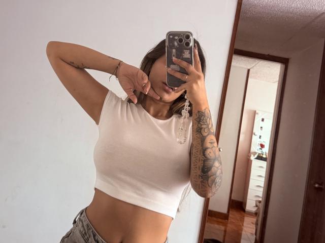 ThaliaAres mit mittelgroßer Brust, in einem weißen Crop-Top, hält ein Telefon, bedeckt ihr Gesicht, zeigt einen tätowierten Arm und trägt graue Hosen, in einem Spiegel-Selfie..