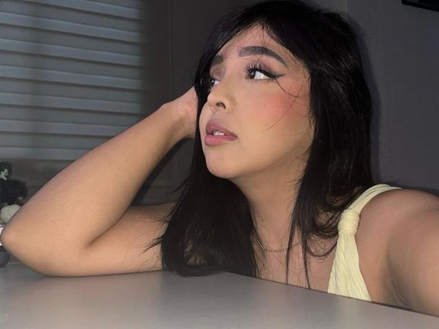 VictoriaKross, avec des cheveux foncés, regardant sur le côté, lèvres roses, eye-liner, une main dans les cheveux, haut jaune, peau claire, collier, assise à une table, éclairage doux, blush discret..