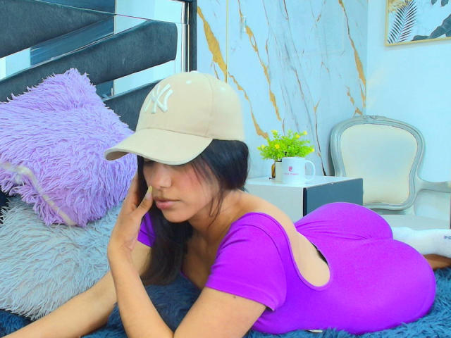 BrianaHarpers, in un body viola, sdraiata a pancia in giù, con i glutei sollevati, indossa un berretto beige, ha i capelli neri e sullo sfondo si vede una sedia bianca..