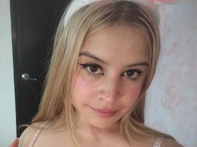 Melani Becker tiene cabello rubio, ojos marrones y una ligera sonrisa. Lleva una blusa rosa de encaje que deja ver parte de su escote, y su rostro está sonrojado con mejillas rosadas..