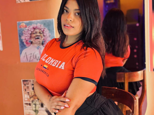 AshlyCarterr with dark hair, orange "BOMBA" shirt, black skirt, crossed arms, wooden chair, mirror reflection, posters on orange wall, subtle smile, medium tits, cute, confident.