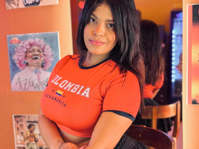 AshlyCarterr with dark hair, orange crop top, showing her big tits, standing with a smile, arms crossed, in a room with colorful posters, reflection in the mirror.