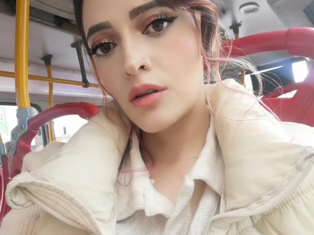 JaneRoa tem pele clara, batom rosa e delineador preto com cílios longos. Ela veste um casaco branco e tem cabelos com mechas rosa, além de um piercing no nariz e a boca ligeiramente aberta..