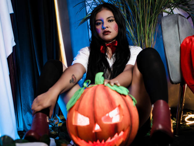 AlessiaVeega with long black hair, white shirt, red bowtie, black knee socks, sitting with legs spread, showing tattoo on left thigh, holding glowing jack-o'-lantern in front of her.