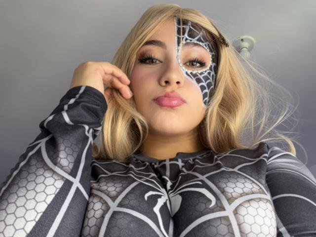 LorensDouce, met blond haar, draagt een strak, zwart en grijs superhelden kostuum met een spinnenweb patroon, dat haar grote borsten, roze lippen en een spinnenweb masker laat zien..