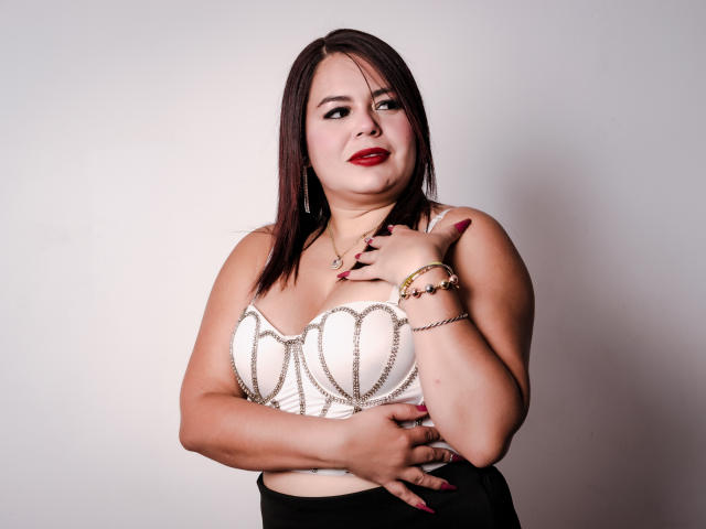 Maya Malik, avec une peau brun clair, du rouge à lèvres, et des cheveux bruns mi-longs, porte un soutien-gorge blanc à motifs de cœur, cachant ses seins avec une main, une jupe noire et un bracelet doré..