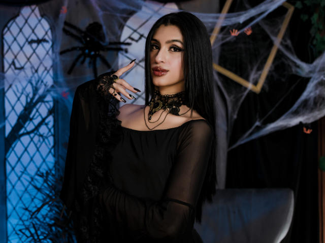 LilithDesire, avec de longs cheveux noirs, un maquillage foncé et un collier en dentelle noire, portant une robe noire transparente, un doigt sur les lèvres, un gant en dentelle noire et des ongles foncés, avec un regard séduisant..