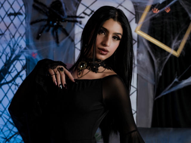 LilithDesire, con i suoi lunghi capelli neri, indossa un abito nero trasparente, una collana e un anello, ha un trucco scuro ed è rivolta verso la fotocamera con un'espressione sexy e misteriosa..