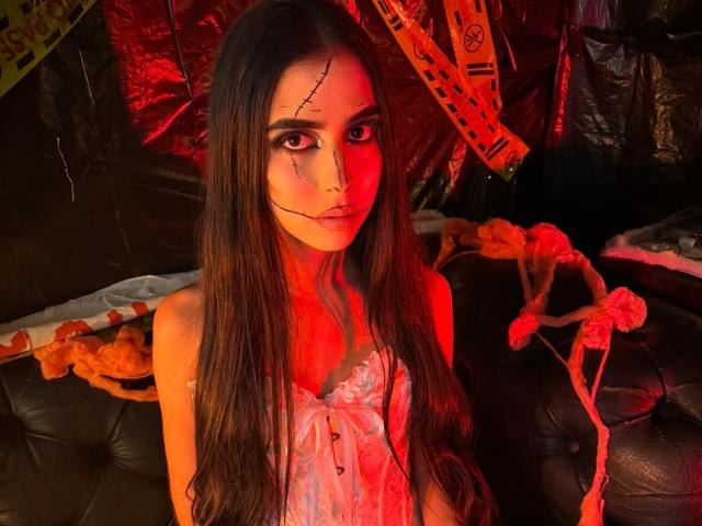 Anna Alvarez, avec ses longs cheveux bruns, son visage illuminé de rouge, son maquillage noir pour les yeux et son haut en dentelle rose, assise sur un canapé en cuir noir, a l'air à la fois sérieuse et séduisante..