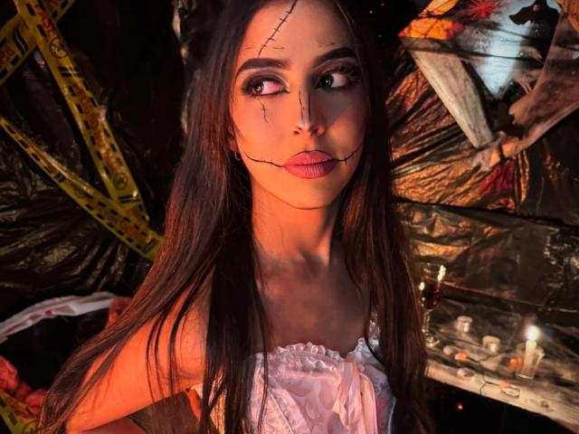 Anna Alvarez, avec de longs cheveux bruns, un maquillage révélant une peau craquelée sur le visage, portant une robe blanche à épaules dénudées, des lèvres roses, regardant vers le haut avec des yeux sombres et expressifs, éclairage orange chaud..