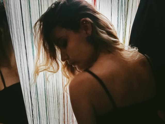 AlanisBlanc with blonde, wavy hair looking down, showing her bare shoulder and black strap, in a dark room with vertical light stripes. Her tits are slightly visible.